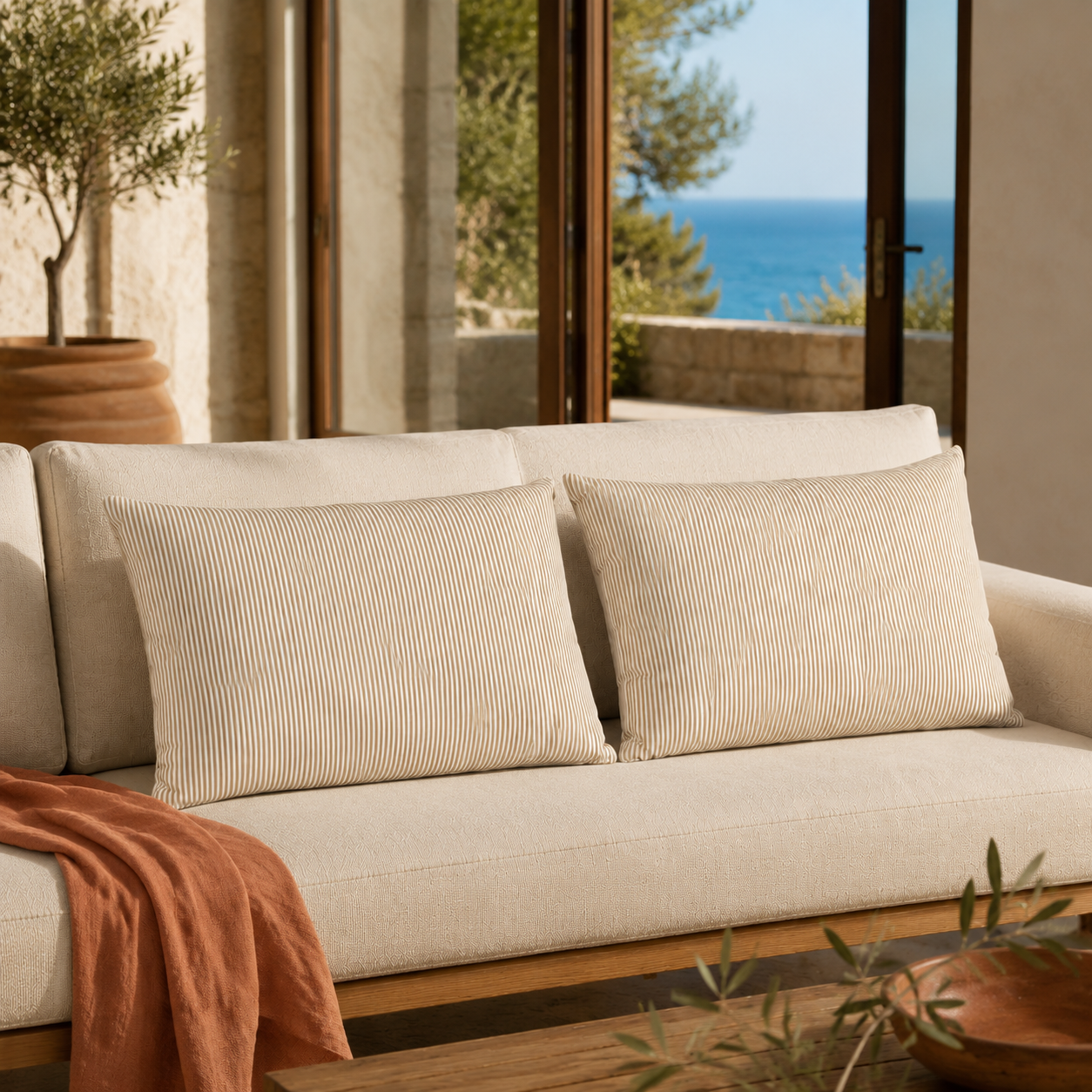 A cream-colored fabric sofa with two matching ribbed throw pillows, styled with a rust-colored throw blanket, set in a sunlit interior with a view of the blue sea through large glass doors.