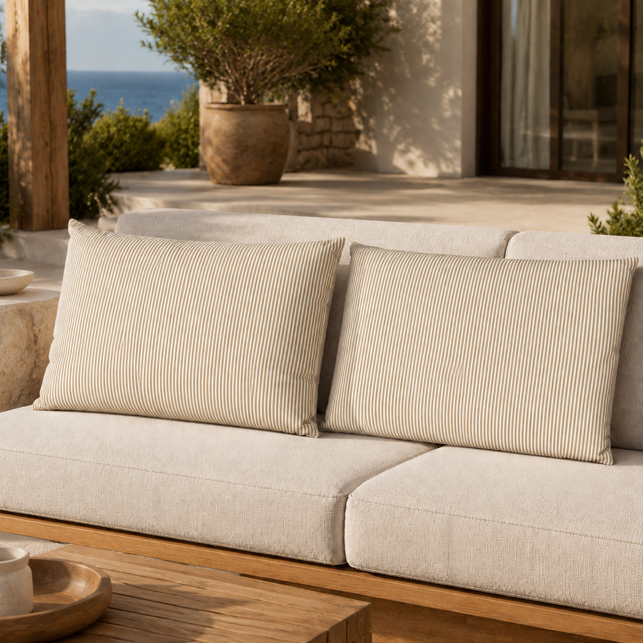 Beige outdoor sofa cushions with thin vertical stripe detailing, shown on a patio with a wooden table and ocean view in the background.