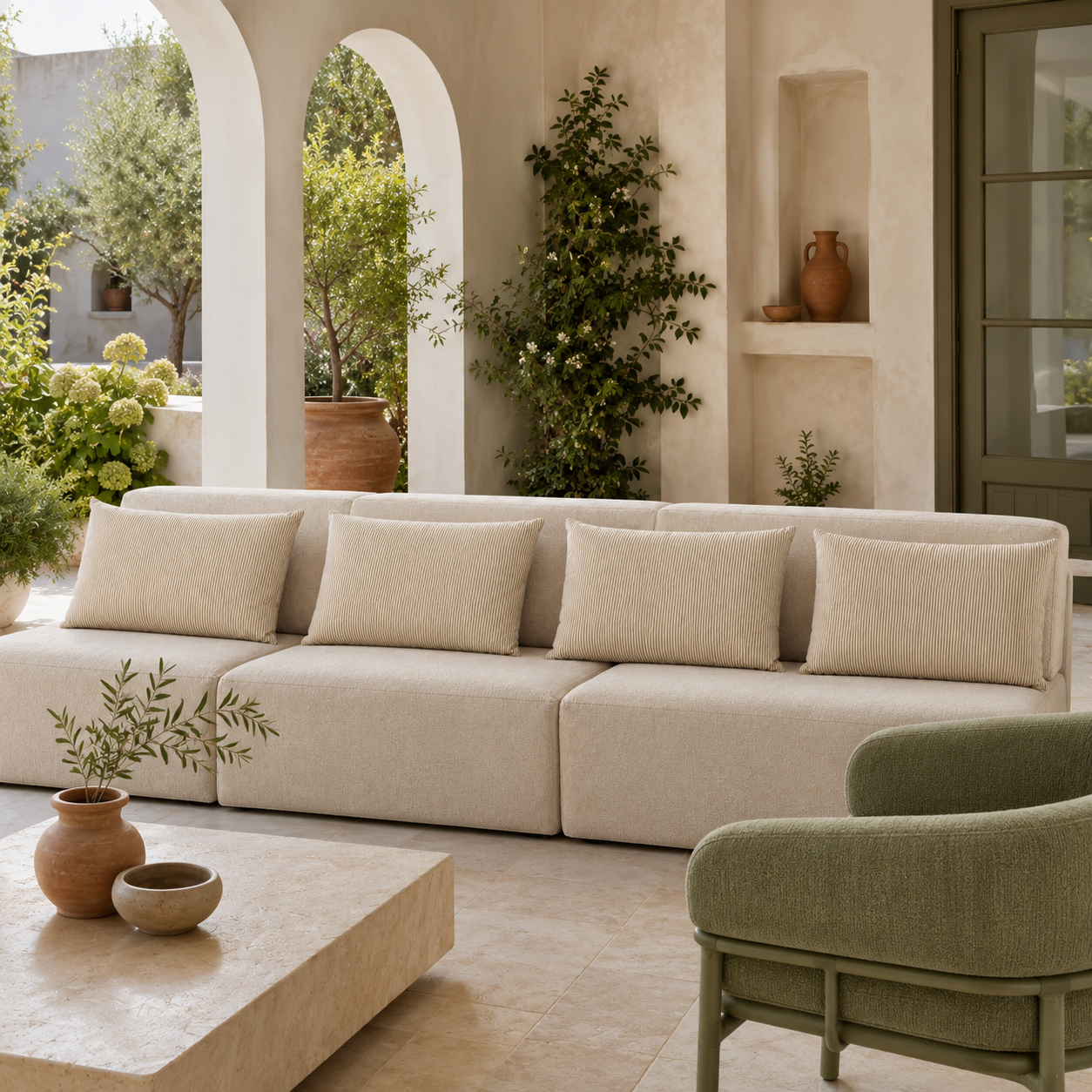 A modern outdoor sectional sofa in a warm cream or light beige bouclé fabric, styled in a Mediterranean patio setting with a matching low coffee table, a sage green accent chair, and terracotta vases.