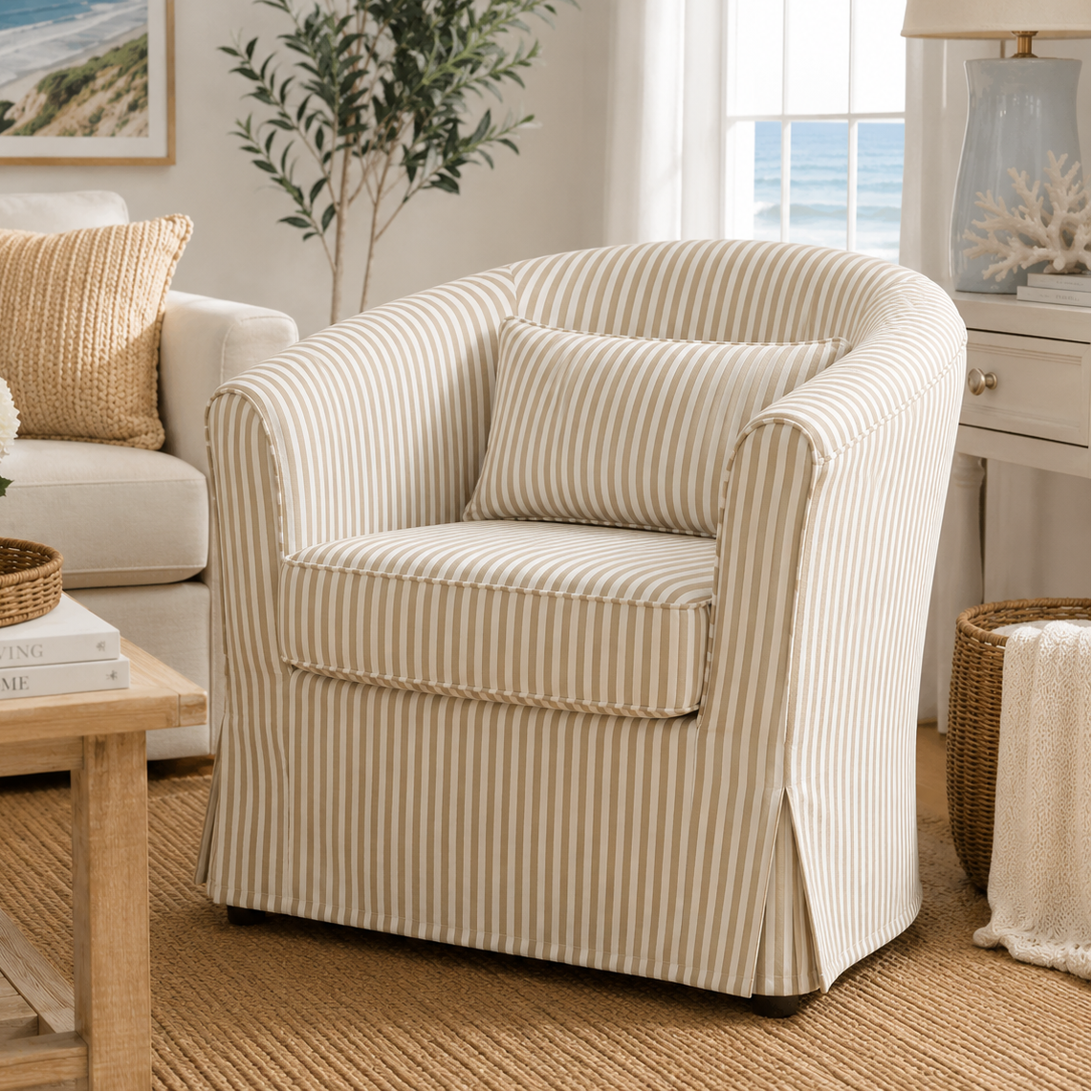 A cream and beige vertically striped slipcovered armchair with a matching throw pillow, styled in a bright, coastal-themed living room next to a wooden coffee table and a white sofa.