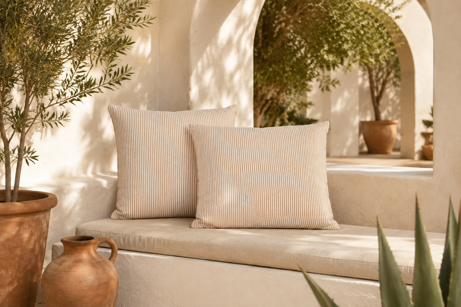 Two beige striped throw pillows resting on a cream cushioned bench in a sunlit Mediterranean-style courtyard with white stucco walls, potted plants, and terracotta vessels.