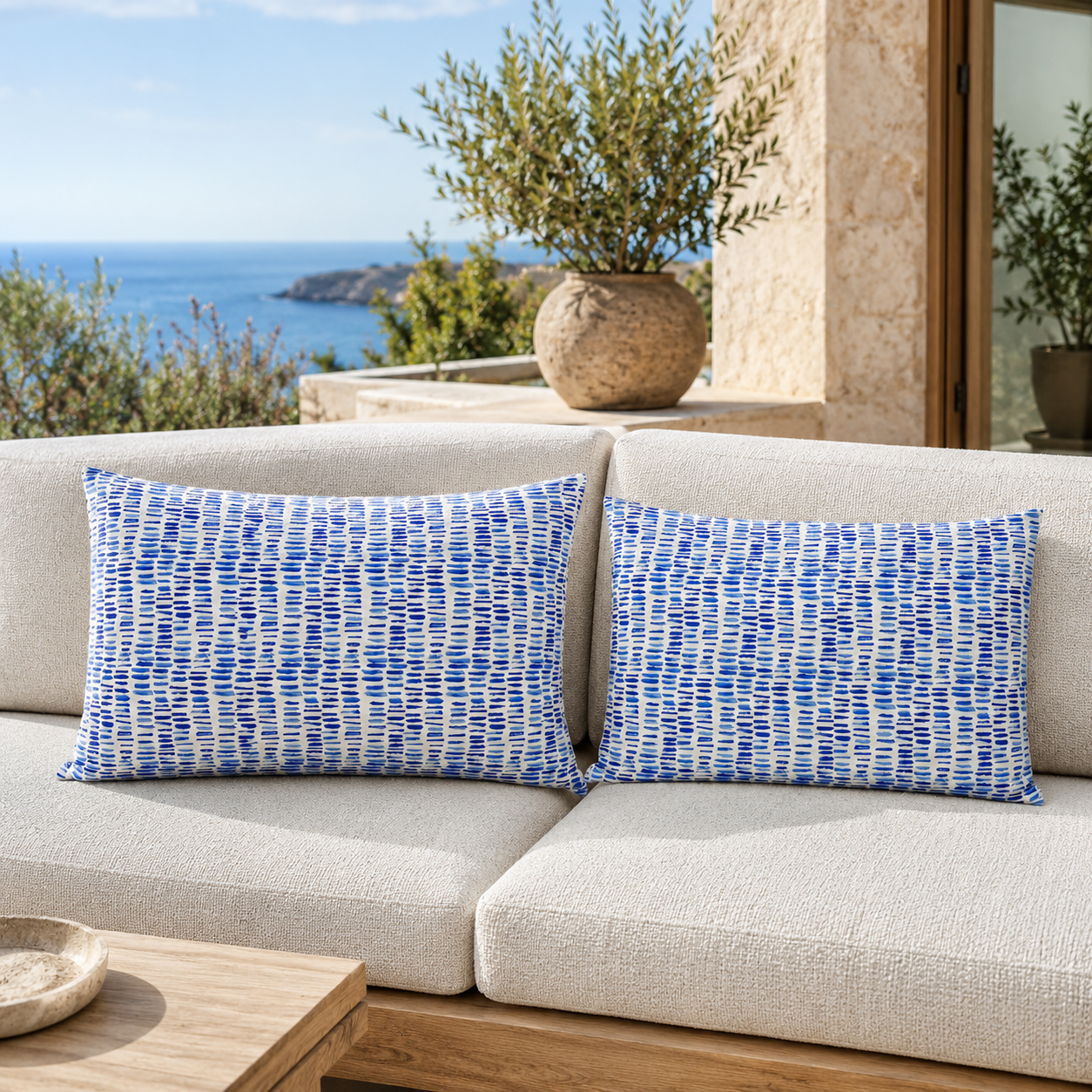 Two rectangular throw pillows with a blue and white textured geometric pattern, styled on a light beige outdoor sofa with a Mediterranean sea view in the background.