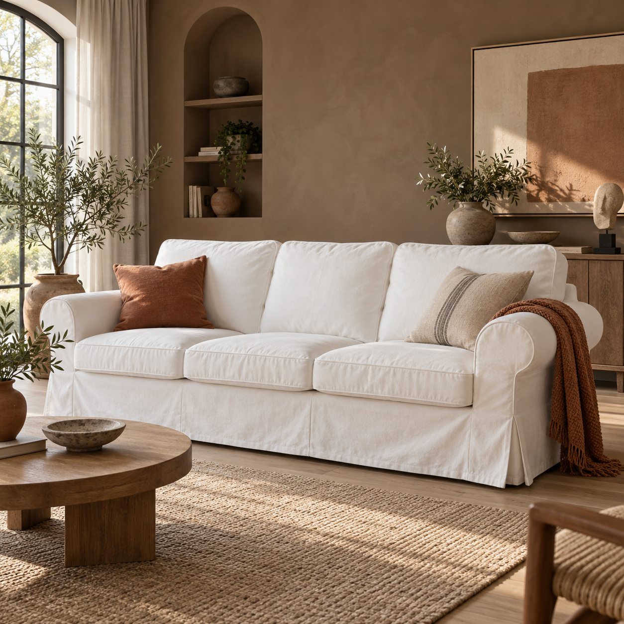 A white slipcovered three-seat sofa in a warm, neutral-toned living room, styled with two decorative throw pillows and a rust-colored throw blanket.