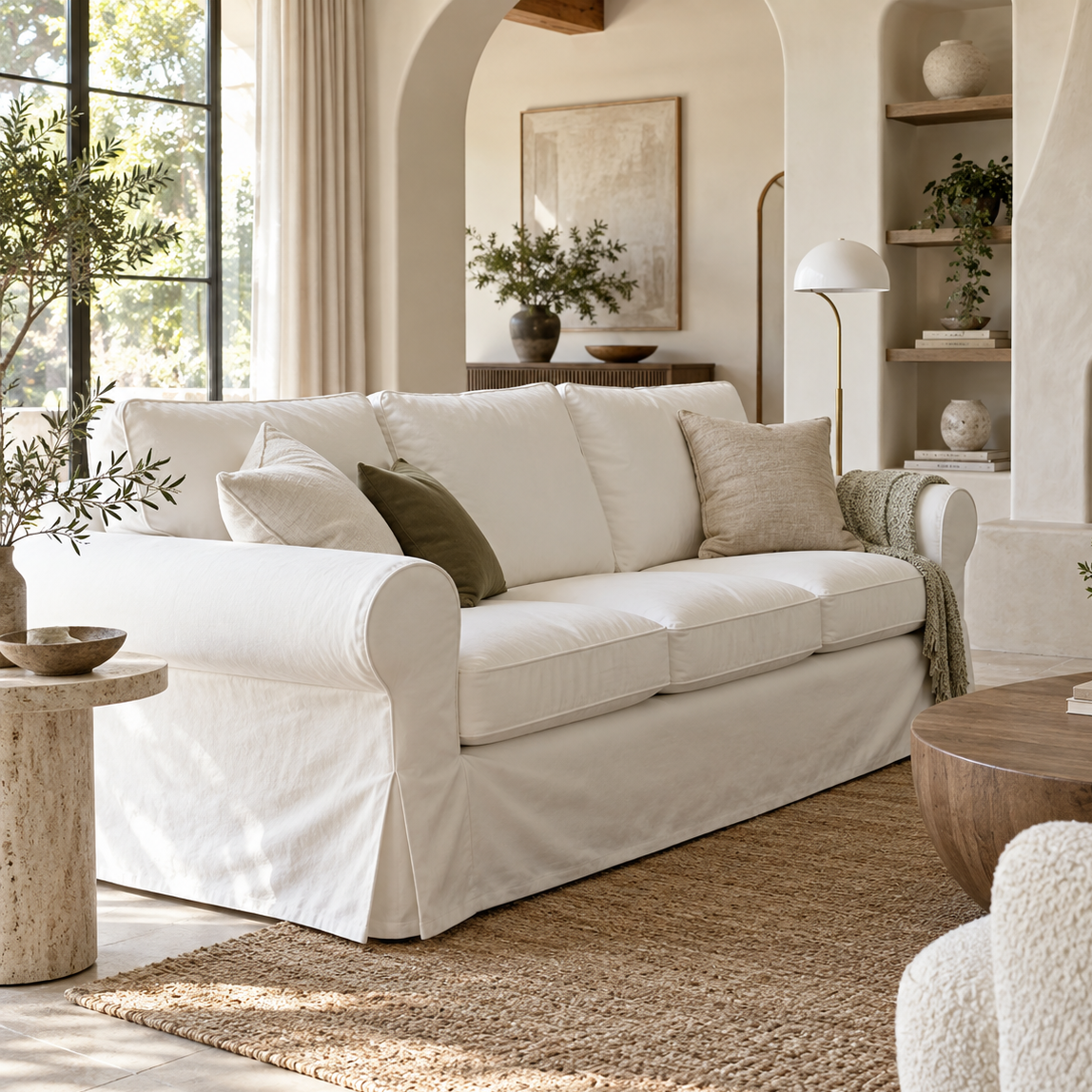 A cream-colored slipcovered sofa in a bright, modern, neutral-toned living room with a woven rug, wooden coffee table, and built-in shelves.