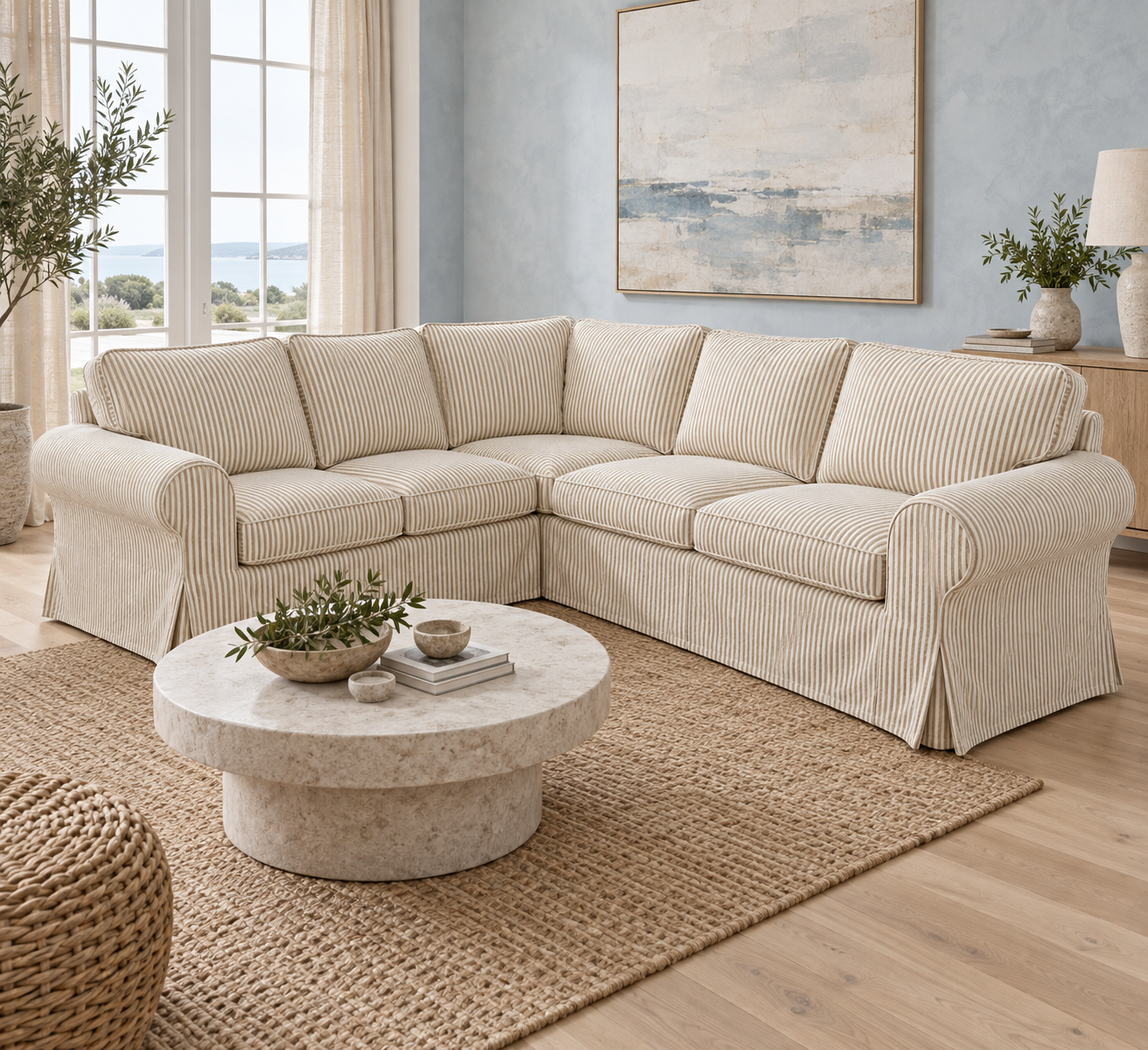 A light beige striped sectional sofa slipcover with matching rolled arms and skirted base, shown in a bright, modern living room setting with a round stone coffee table and a jute rug.
