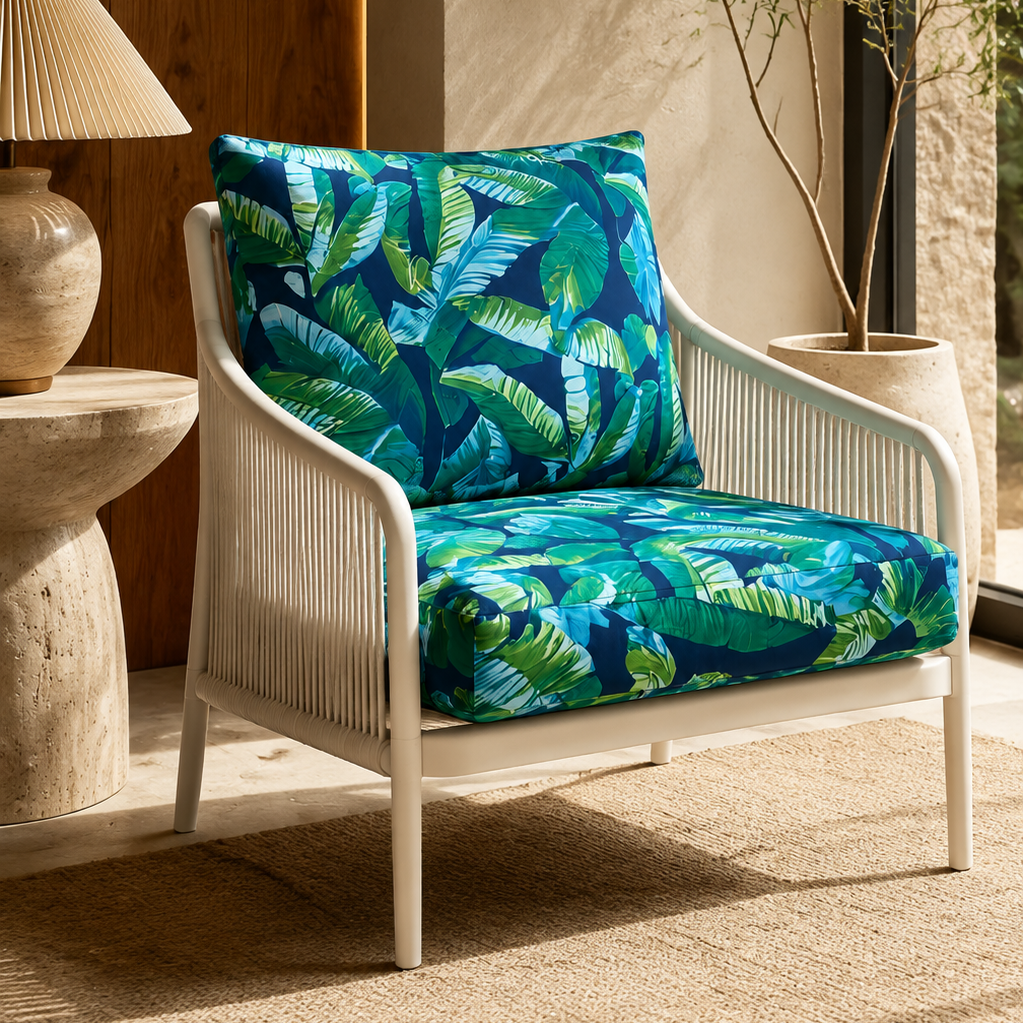 A white outdoor lounge chair with a tropical leaf print cushion set, styled in a warm, sunlit patio setting next to a side table with a lamp and a potted plant.