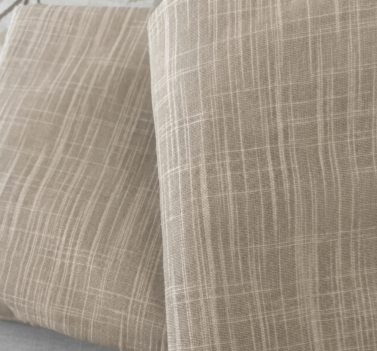 A close-up of folded fabric with a fine plaid pattern in warm beige and taupe tones, showing the woven texture clearly.