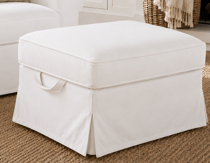 A square storage ottoman upholstered in off-white fabric with a skirted base and a fabric carry handle, shown in a bright, neutral room setting on a woven rug.