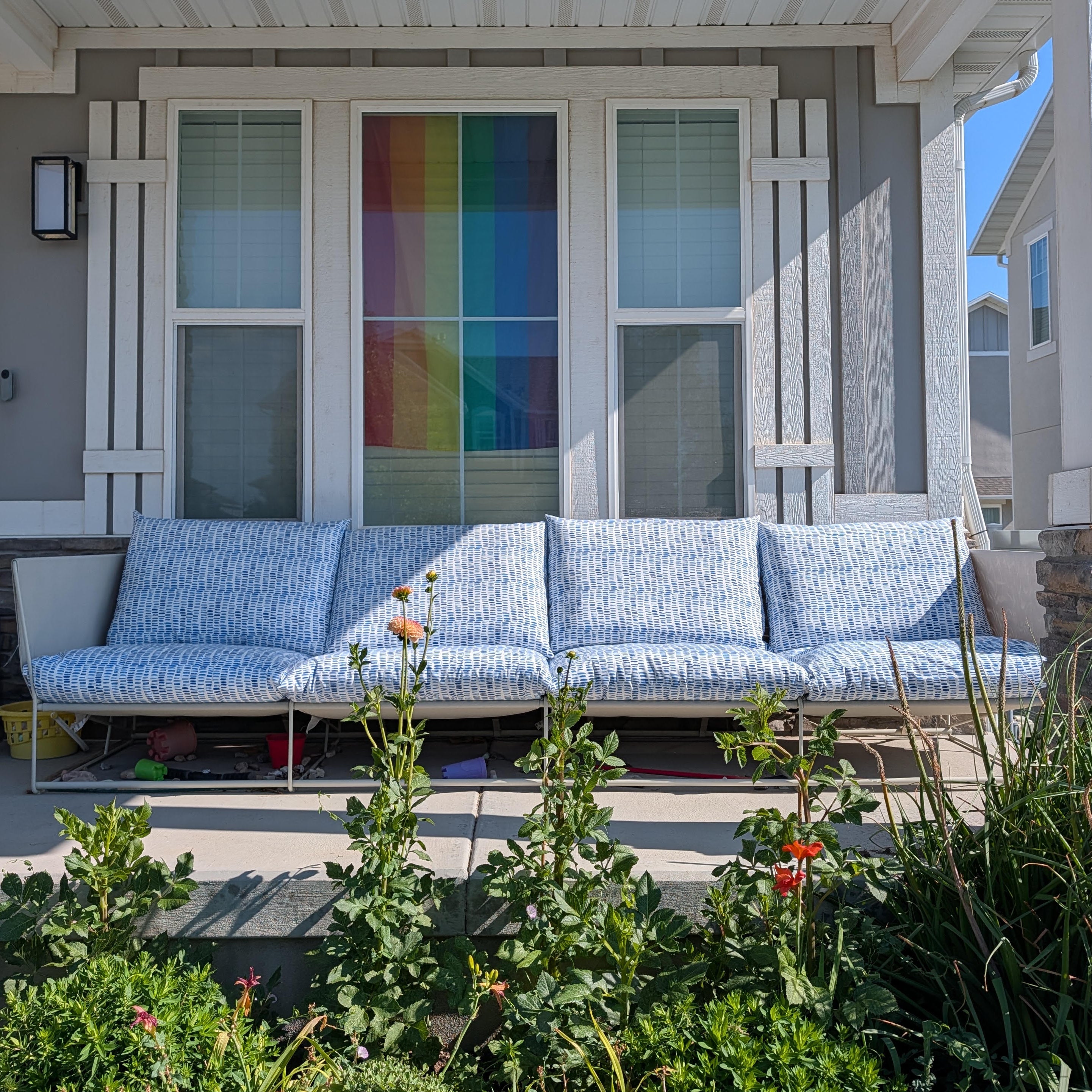 A modern outdoor daybed with light blue and white patterned cushions sits on a covered porch in front of a gray house with white trim and a colorful rainbow window pane. Green garden plants are visible in the foreground.