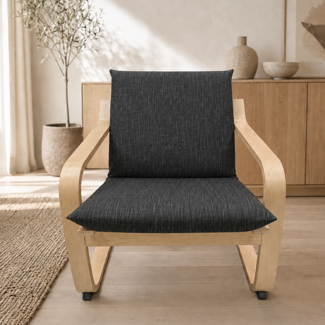 A modern wooden armchair with a curved light oak frame and dark charcoal textured seat and back cushions, shown in a bright, minimalist room with a wooden cabinet and potted plant in the background.