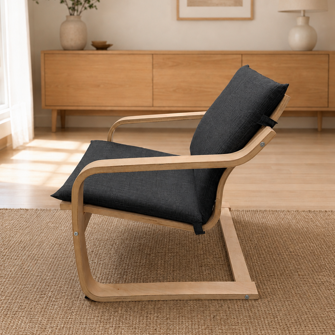 A modern lounge chair with a light natural wood frame and dark charcoal gray fabric cushions, placed on a woven jute rug in a bright, minimalist living room.