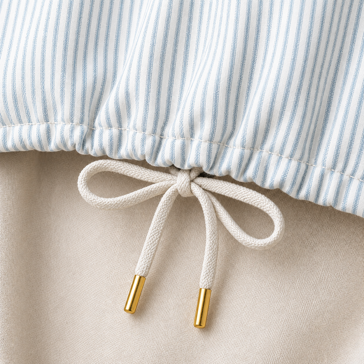 Close-up of a drawstring bag or pouch with a light blue and white striped fabric top, a cream-colored drawstring tied in a bow, and gold metal tips, set against a neutral beige background.
