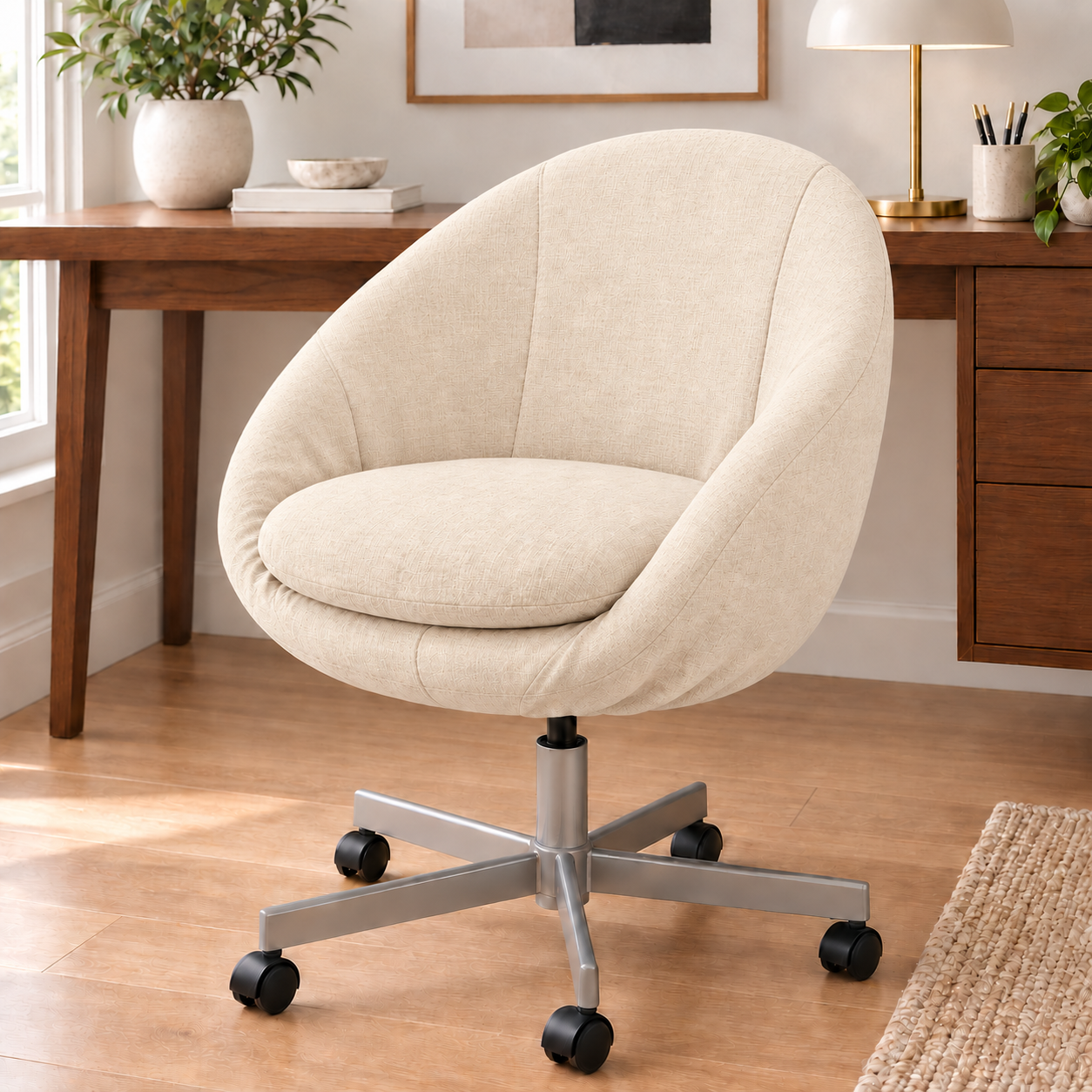 A cream-colored fabric swivel office chair with a silver five-star base and black casters, set in a bright home office next to a wooden desk.