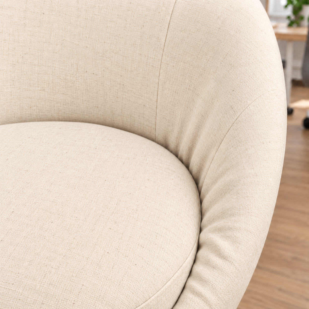 Close-up of a curved upholstered chair in a light beige textured fabric, showing the seat cushion and rounded backrest in a softly lit indoor setting with a blurred wood floor and furniture in the background.