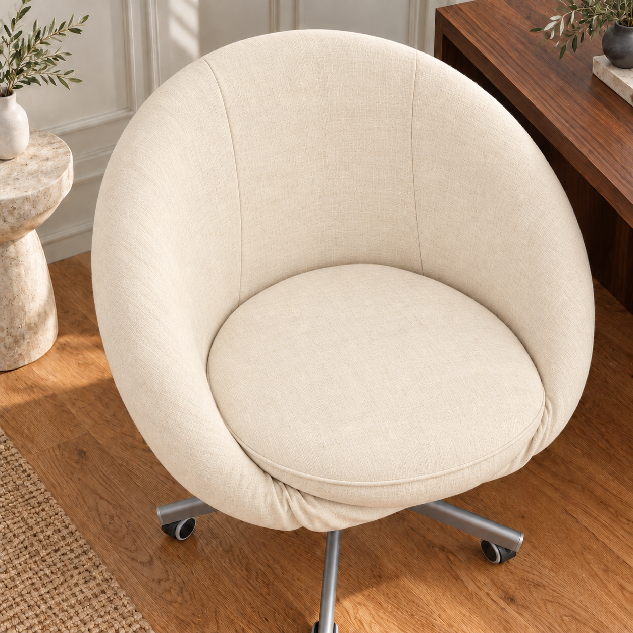 A cream-colored fabric swivel office chair with a round cushioned seat and backrest, mounted on a silver metal five-star wheeled base, shown in a home office setting.