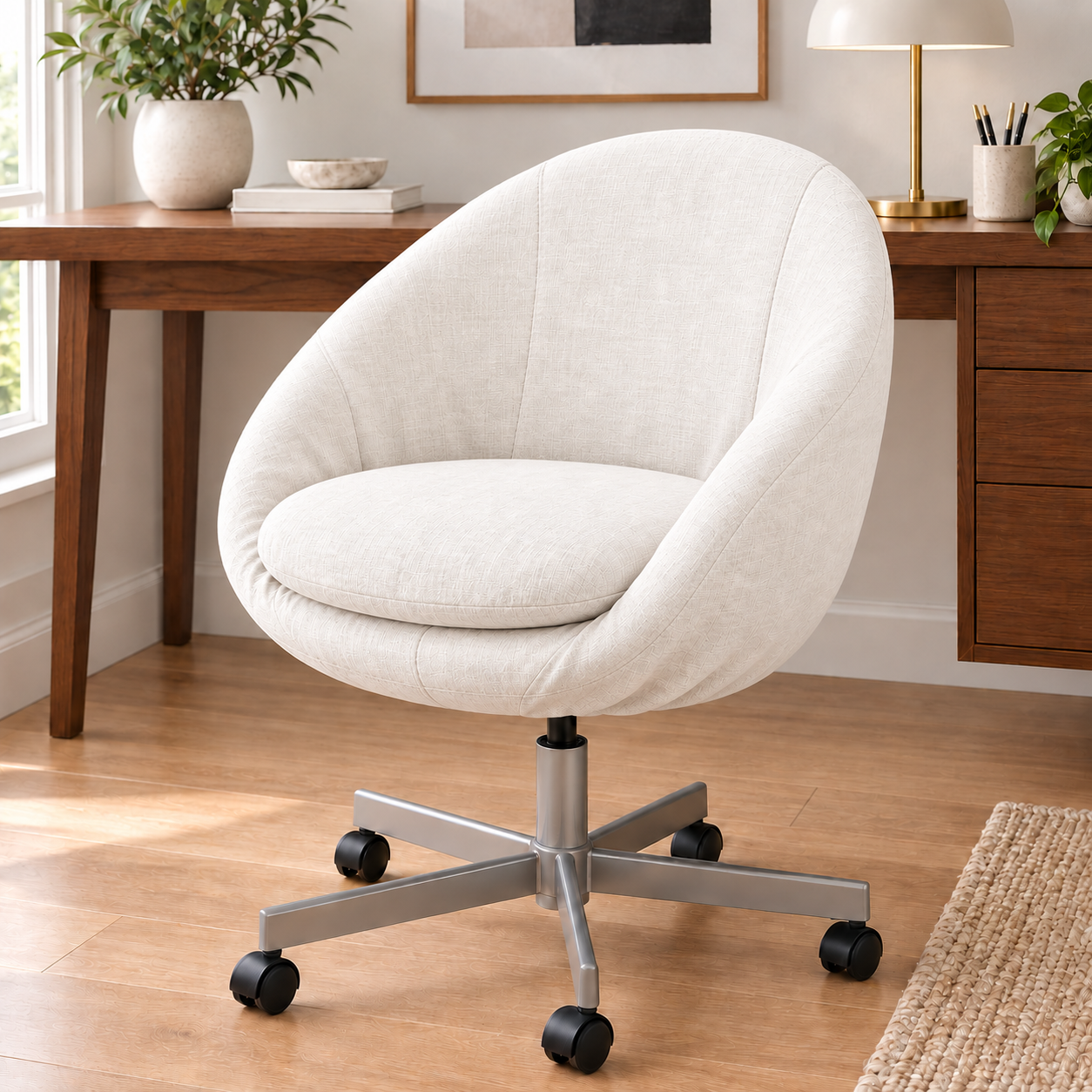 A modern upholstered swivel office chair with a rounded backrest and seat cushion in off-white fabric, mounted on a silver metal five-star base with black casters, shown in a home office setting.