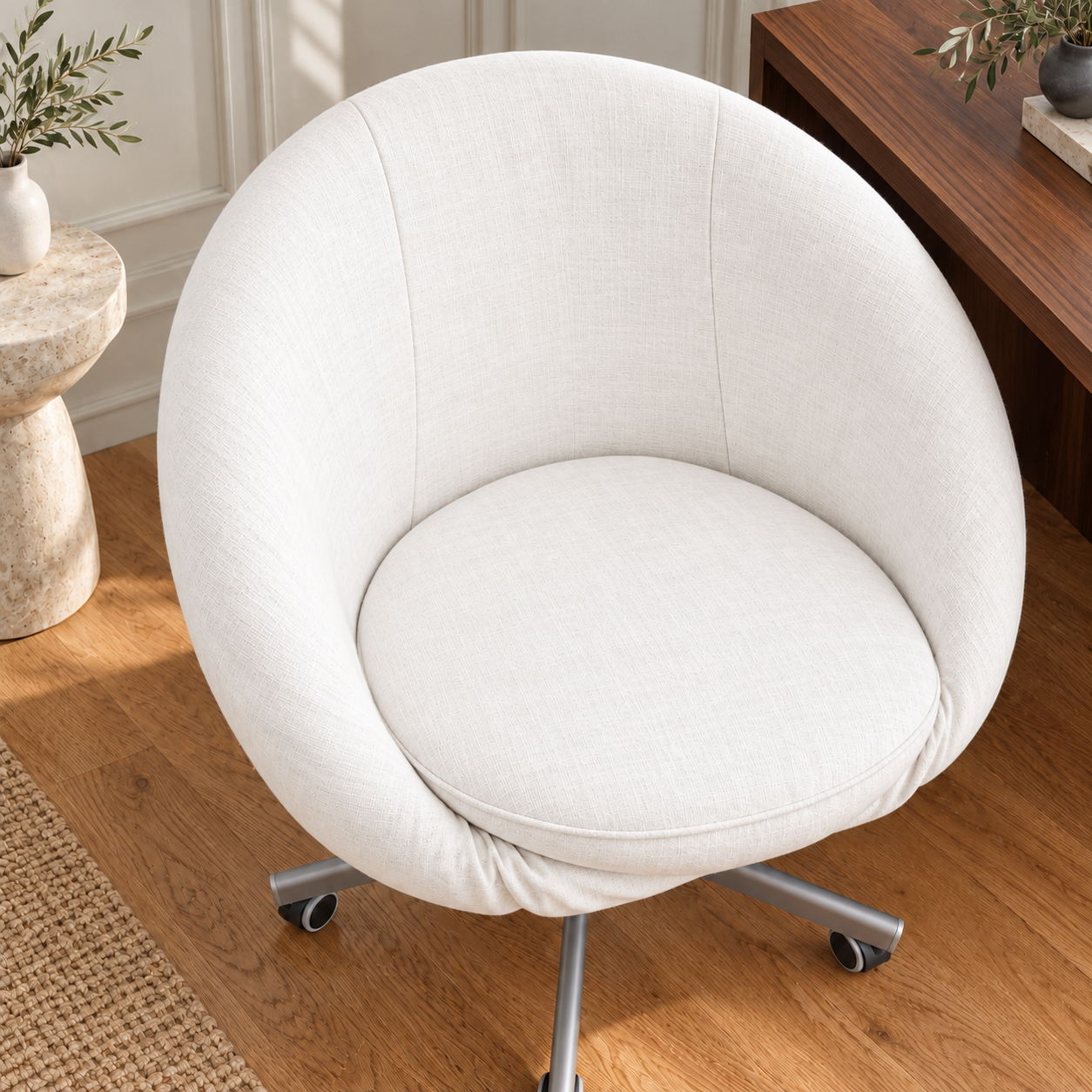 A modern round upholstered swivel chair with a cushioned seat and backrest, shown in a warm indoor room with wood flooring and a wooden desk in the background.