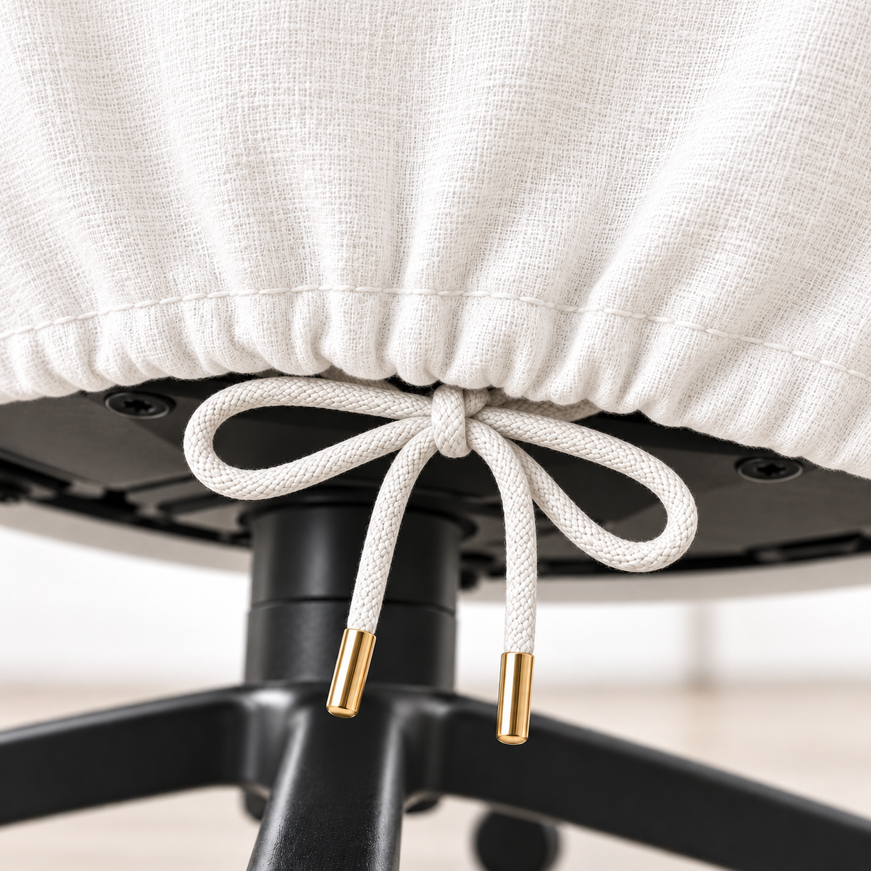 Close-up of a white textured fabric chair slipcover tied under the seat with a white rope drawstring and gold metal tips, attached to a black office chair base.