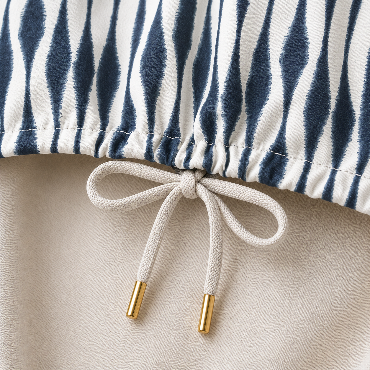 Close-up of a drawstring garment detail showing a navy and white vertically striped fabric at the top, a cream drawstring tied in a bow at the center, and gold-toned metal tips on a light beige fabric below.