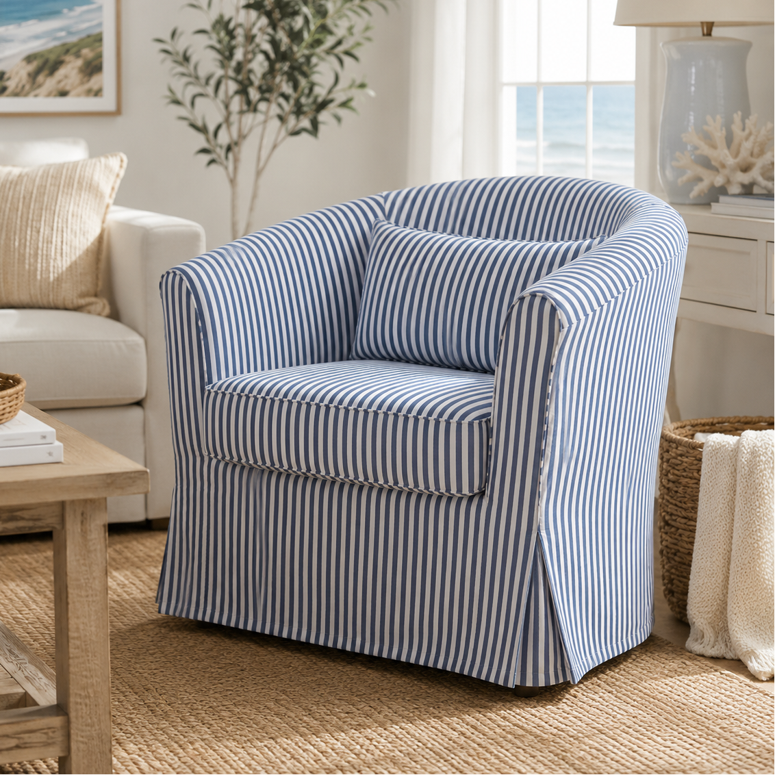 A slipcovered barrel-style armchair with blue and white vertical stripes, shown in a bright, coastal-themed living room next to a white sofa and a wooden coffee table.