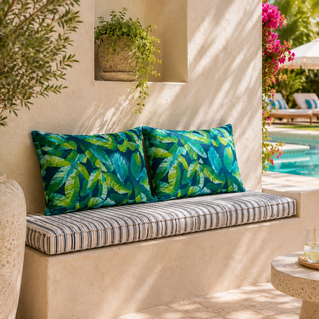 Two tropical leaf print throw pillows and a striped cushion on a built-in outdoor bench beside a pool, with palm trees, lounge chairs, and a patio setting in the background.