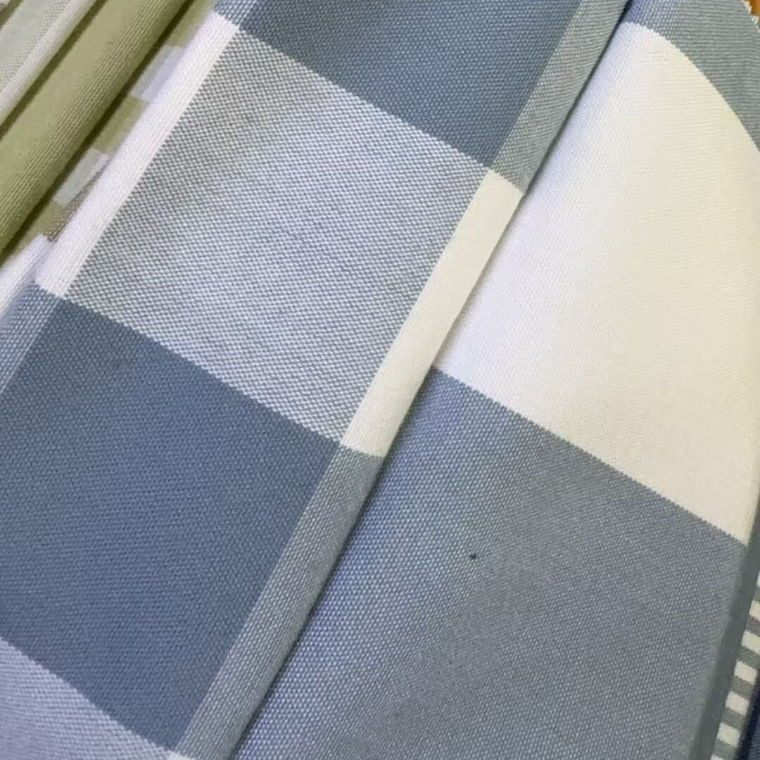 A close-up of folded fabric with a large blue and white check pattern, alongside other folded textiles in muted green, gray, and striped designs.