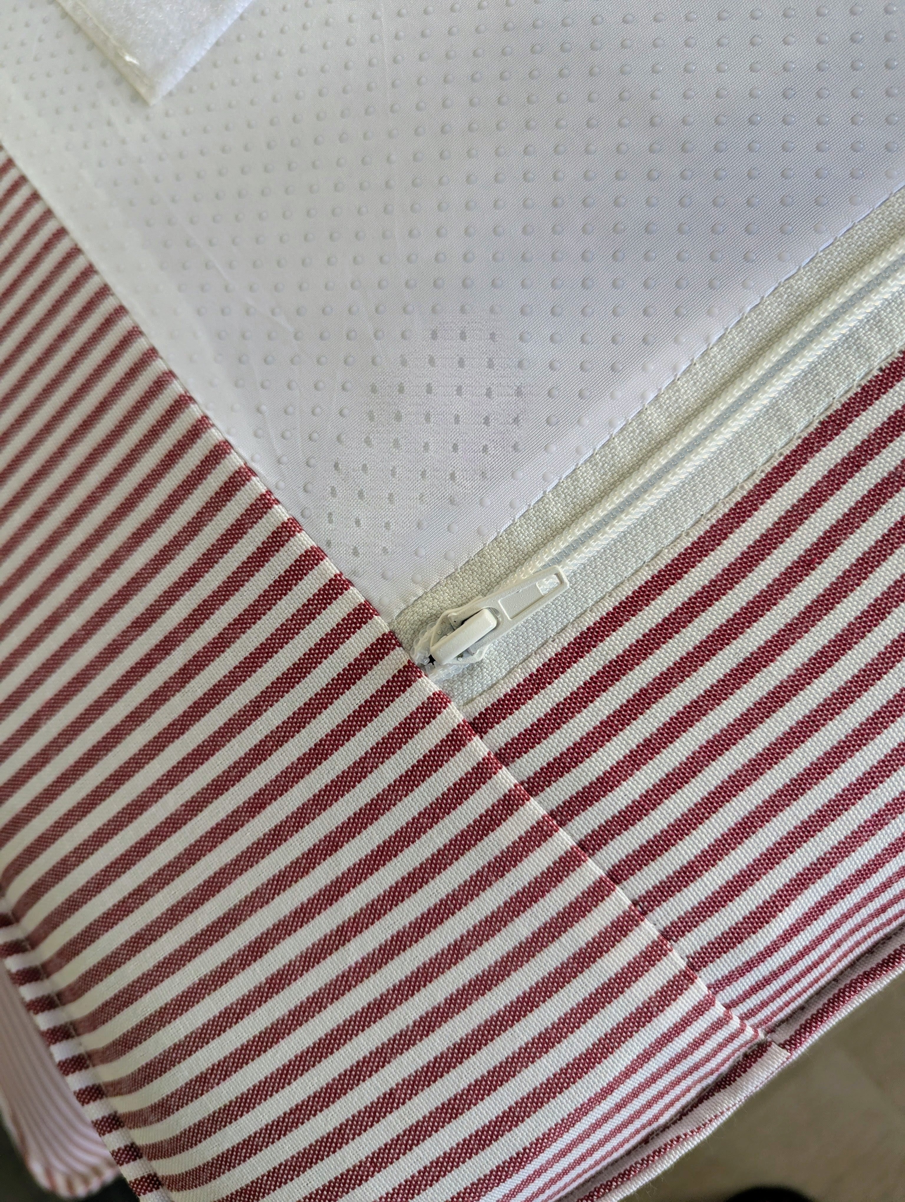 Close-up of a striped fabric item, likely a bed skirt or fitted sheet, with terracotta red and off-white horizontal stripes and a white zipper opening to a white mesh insert.
